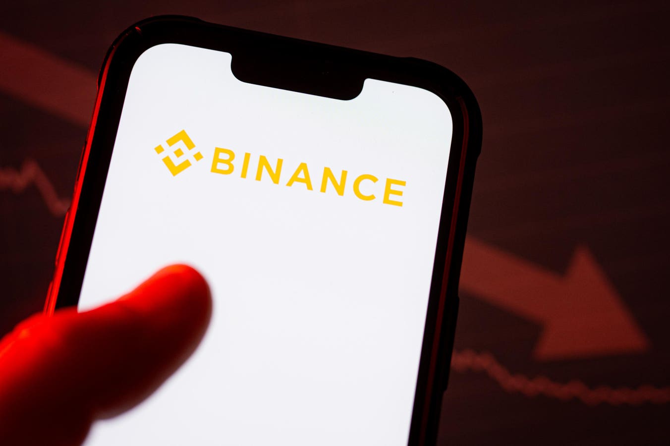Mão diante de um celular com o logotipo da Binance na tela, com um gráfico em queda ao fundo