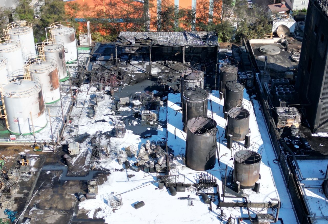 Vista aérea de tanques de armazenamento queimados numa fábrica de produtos químicos, com espuma branca no chão e tanques intactos ao lado