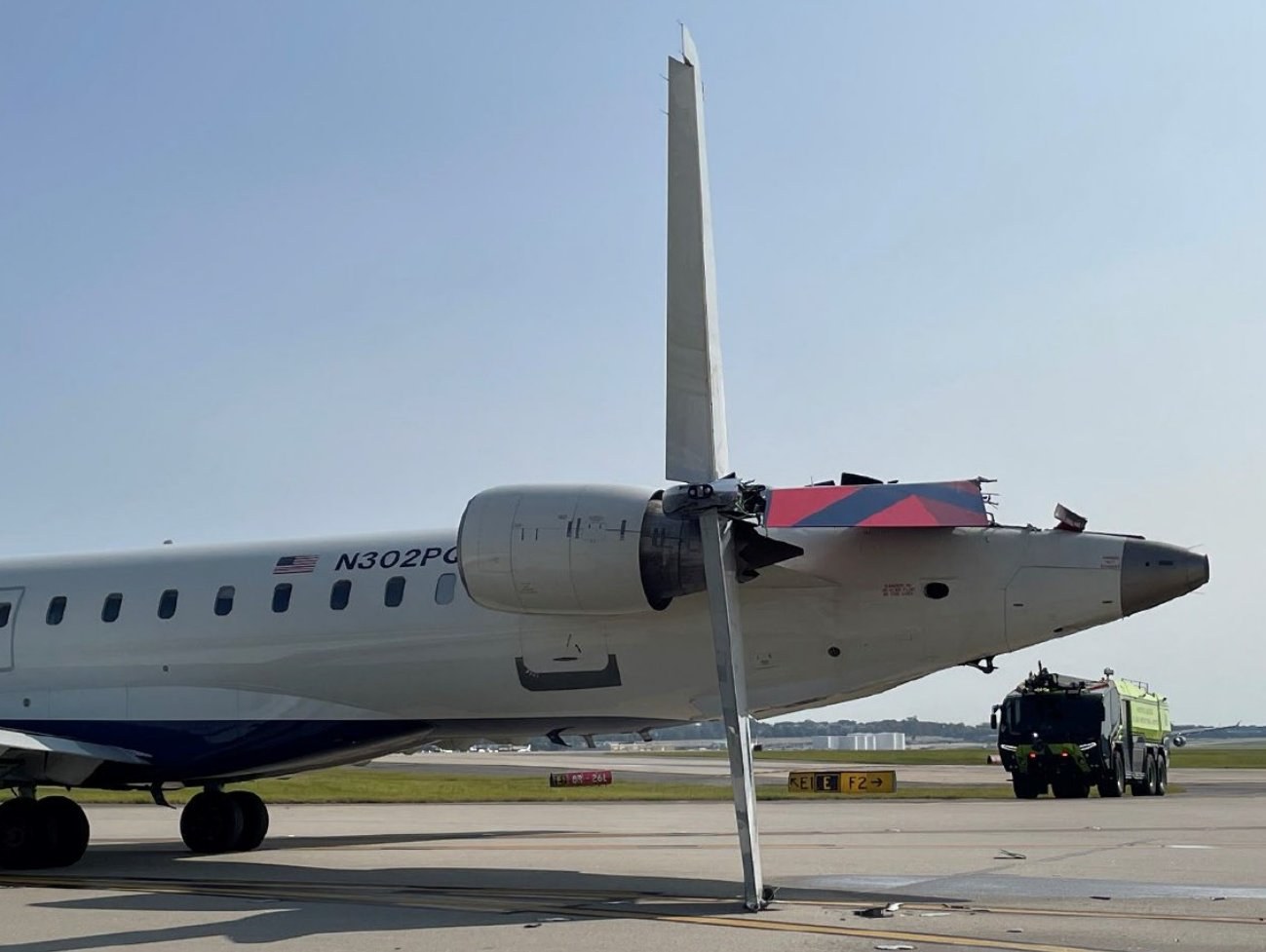 Avião regional CRJ-900 visto de lado, com a parte traseira danificada e o estabilizador vertical deformado, com um caminhão de bombeiros ao fundo