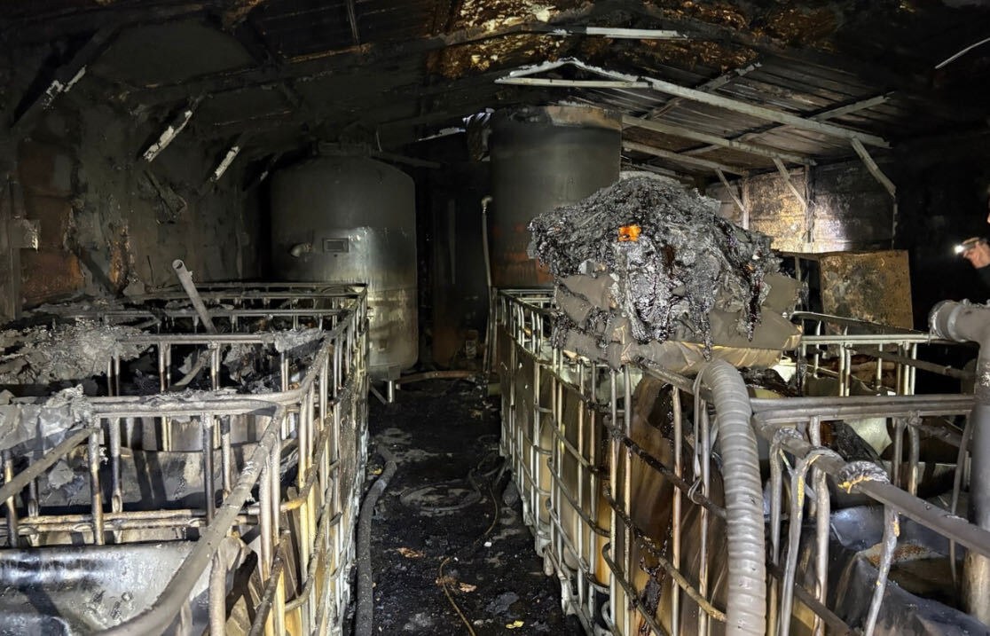 Interior queimado do galpão da destilaria, com tanques metálicos e contêineres de armazenamento carbonizados