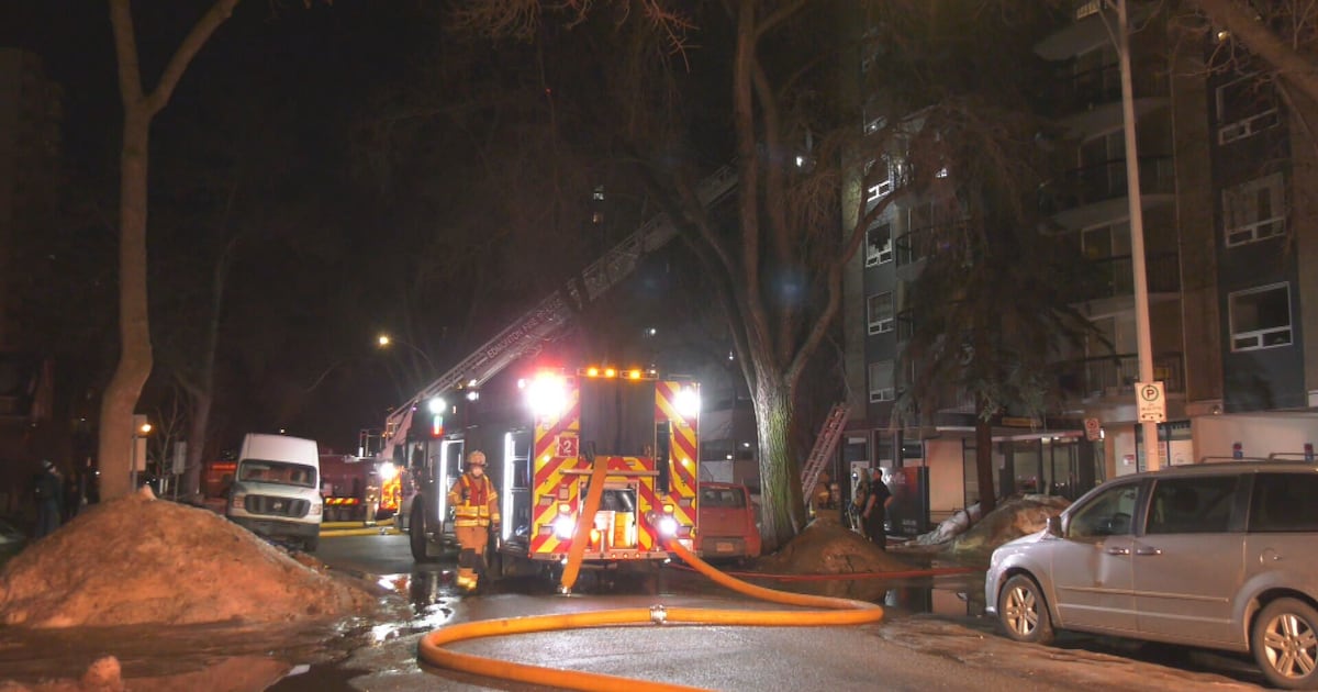 Caminhão de bombeiros com escada estendida e mangueira na rua, à noite, diante de um prédio residencial
