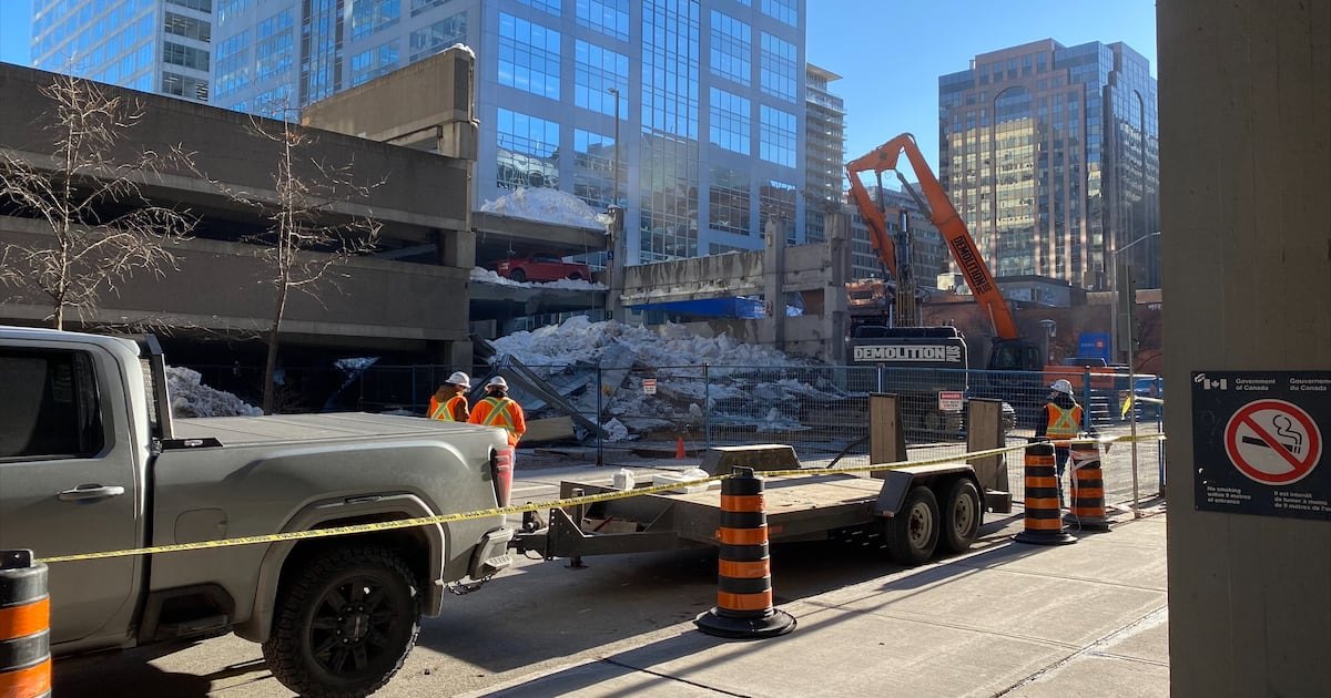 Equipes de demolição e uma escavadeira junto ao edifício-garagem parcialmente desabado no centro de Ottawa, com neve e entulho atrás de tapumes