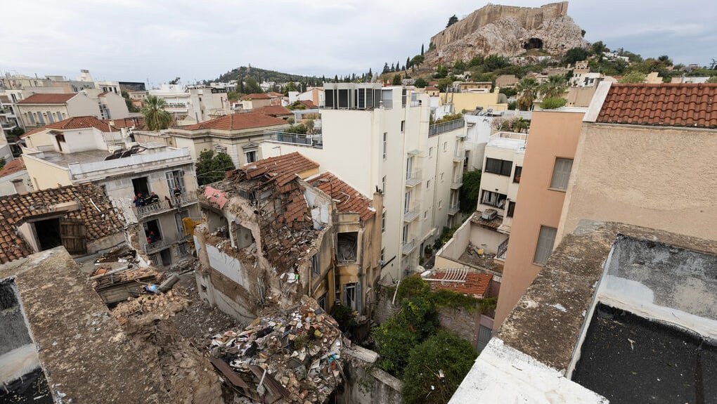 Prédio desabado entre casas do bairro de Plaka, em Atenas, com a Acrópole ao fundo e equipes de resgate trabalhando nos escombros