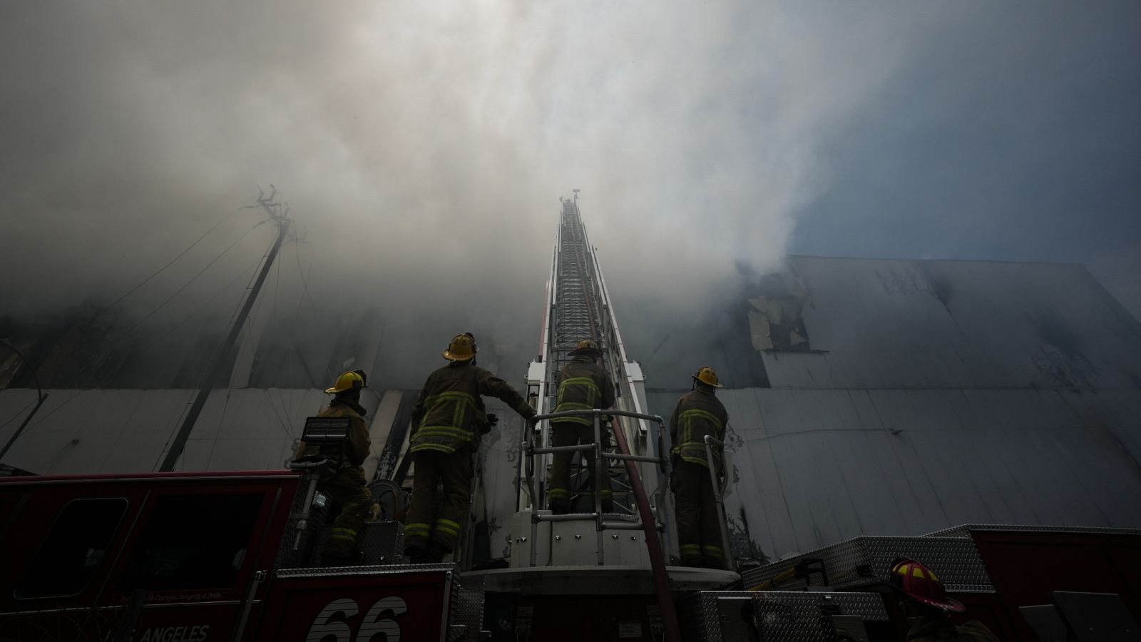 Bombeiros sobre um caminhão com escada aérea estendida diante da fachada de um armazém tomado por fumaça