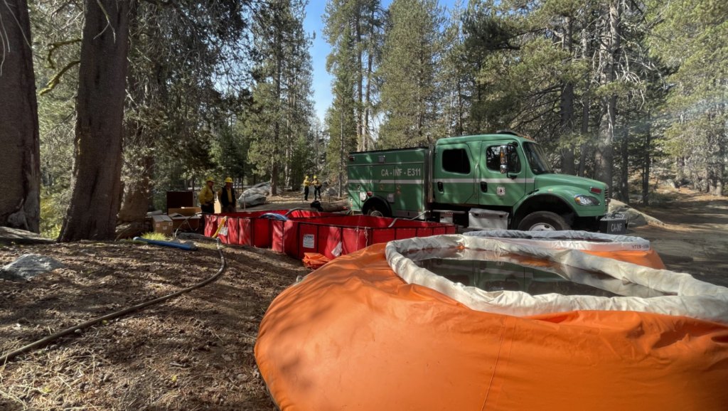 Caminhão verde de combate a incêndio florestal ao lado de reservatórios portáteis de água, vermelhos e laranja, numa área de pinheiros