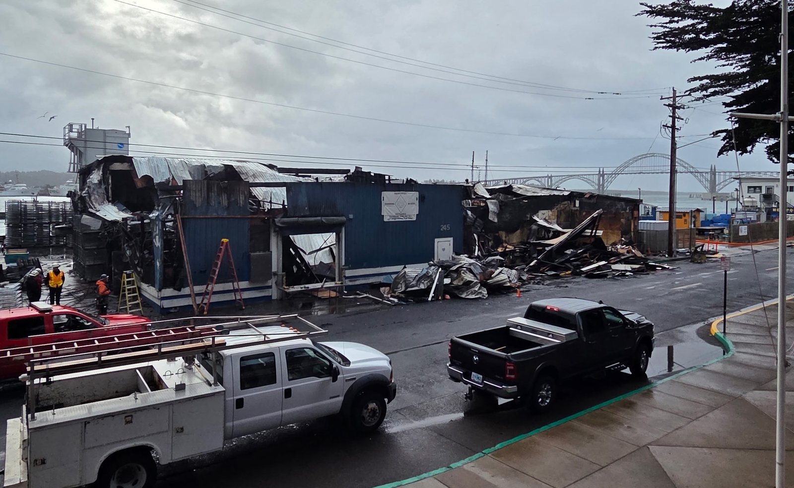 Prédio frigorífico da Pacific Seafood destruído pelo fogo na orla de Newport, com escombros na rua, caminhonetes estacionadas e a ponte da baía ao fundo