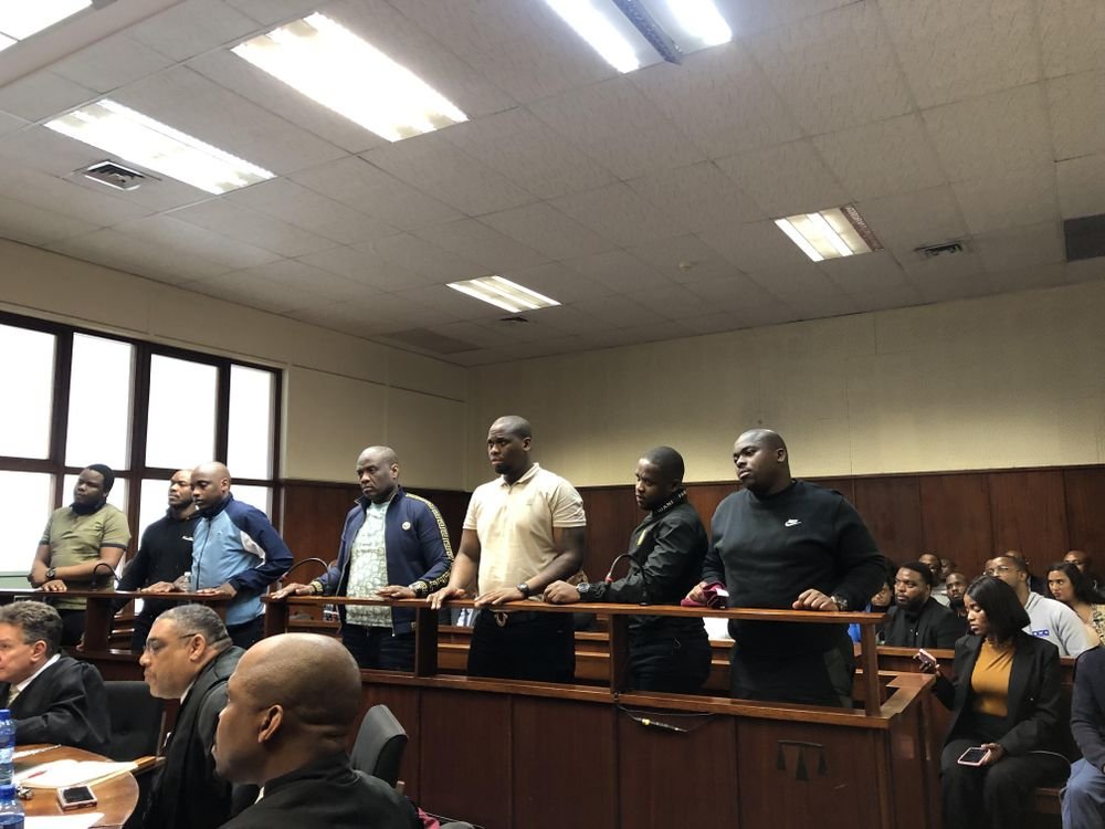 Os sete acusados de pé no banco dos réus da sala de audiências em Durban, com advogados à frente e público ao fundo