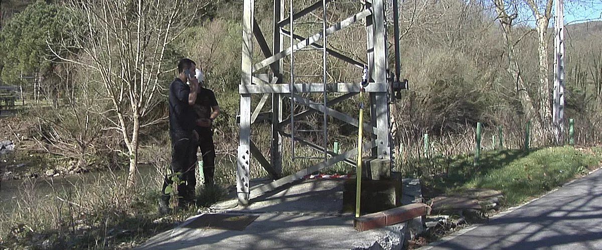 Base de uma torre metálica de alta tensão com escada vertical, ao lado de uma via de pedestres, com duas pessoas junto à estrutura