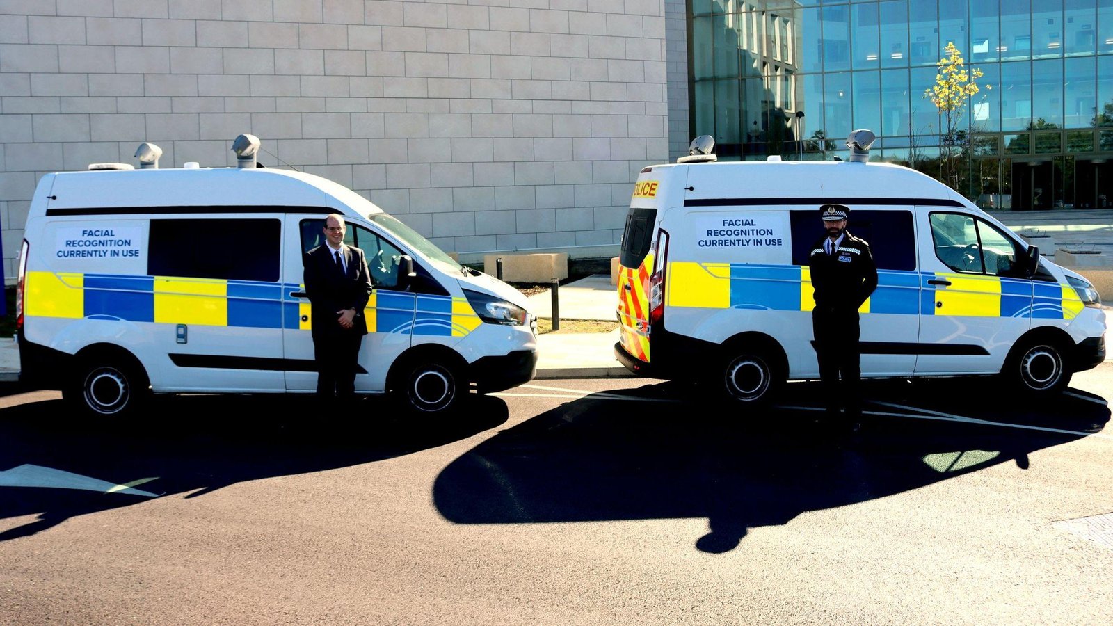 Duas vans brancas da polícia com faixas azuis e amarelas e o aviso "Facial Recognition Currently In Use" (reconhecimento facial em uso), com um homem de terno e um policial fardado ao lado