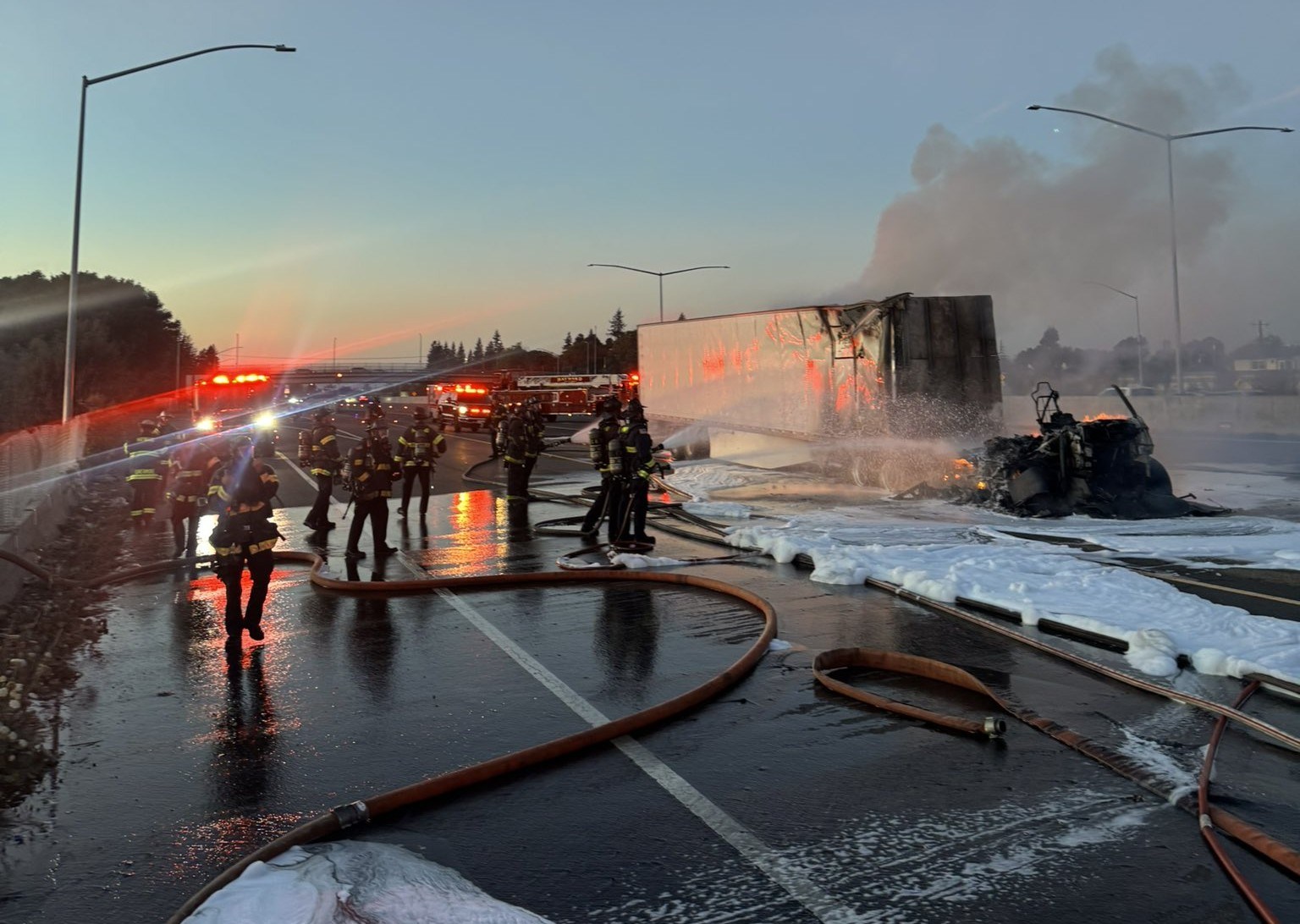 Bombeiros com mangueiras e espuma ao lado da cabine queimada de um caminhão e de um reboque chamuscado numa rodovia ao entardecer