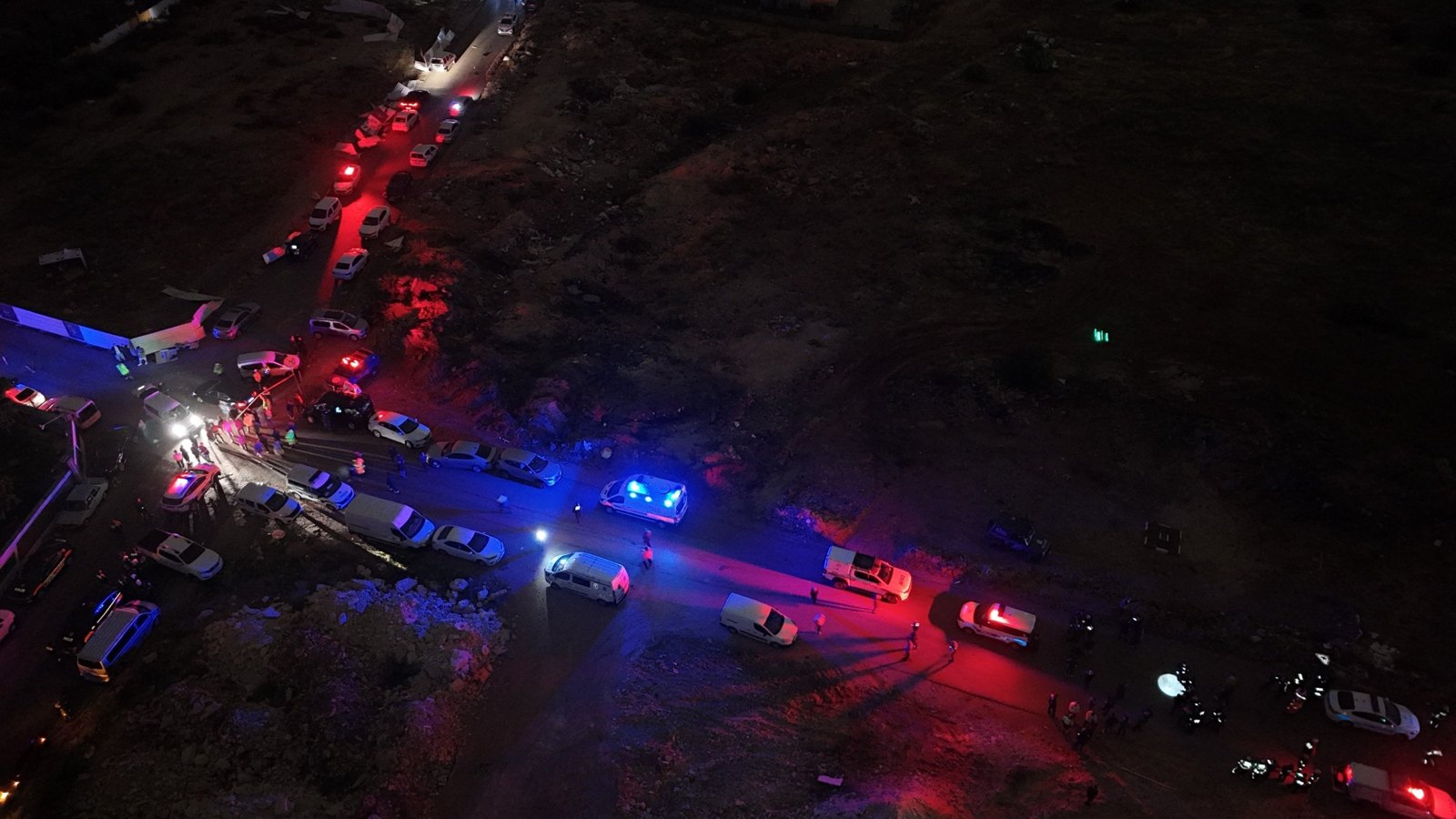 Vista aérea noturna do local do tornado em Antalya, com viaturas de polícia, ambulância e equipes de resgate enfileiradas numa rua de terra ao lado do canteiro
