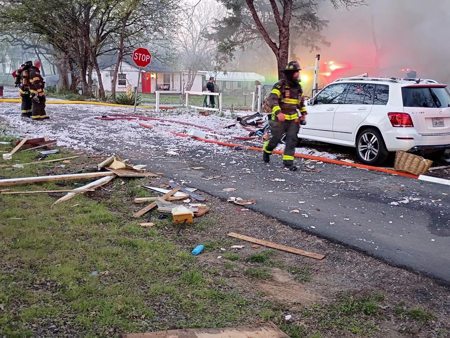 Bombeiros caminham por uma rua residencial coberta de destroços e fragmentos de material isolante, com fumaça e viaturas ao fundo