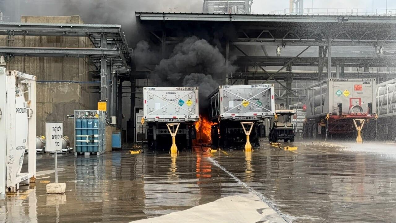 Fumaça preta e fogo entre dois reboques de cilindros de gás estacionados sob a cobertura de uma área industrial molhada
